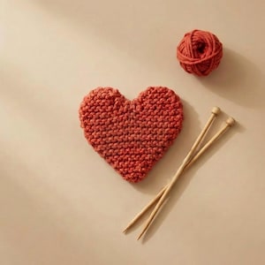 May include: A knitted heart in a reddish-orange colour, a ball of matching yarn, and two wooden knitting needles are arranged on a neutral background. The heart-shaped item is the focal point, suggesting a handmade craft project.