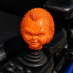 May include: A bright orange, molded plastic doll head featuring a textured hairstyle and visible stitched scar details across the forehead and cheeks, mounted as a decorative joystick topper on a black control panel with circular buttons.