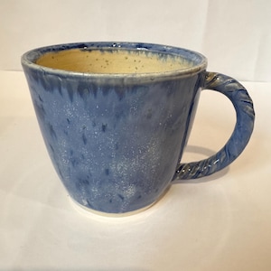 Puede incluir: Una taza de cerámica azul con interior blanco y asa en forma de cuerda. La taza tiene un esmalte moteado.