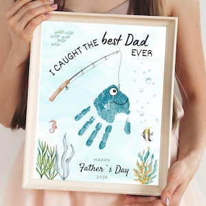 Puede incluir: Impresión enmarcada con un marco de madera clara. La obra de arte presenta una huella de mano azul con forma de pez, con una caña de pescar y el texto "I caught the best Dad ever". El texto adicional dice "Happy Father's Day 2026".
