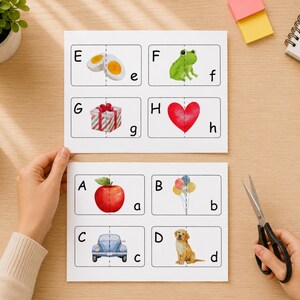 May include: Two white sheets of paper featuring educational alphabet matching cards with uppercase and lowercase letters paired with illustrations of an egg, frog, gift, heart, apple, balloons, car, and dog, each divided by a dashed cutting line.