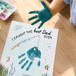 May include: A white rectangular paper featuring a blue handprint transformed into a fish with a drawn eye and smile. The text reads I CAUGHT THE best Dad EVER. A fishing rod illustration is positioned above the fish, surrounded by small sea creatures.