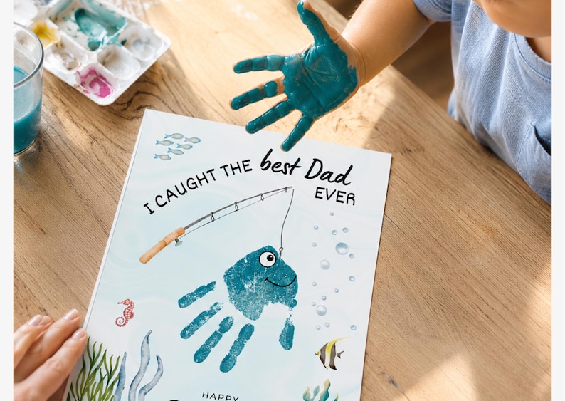 May include: A white rectangular paper featuring a blue handprint transformed into a fish with a drawn eye and smile. The text reads I CAUGHT THE best Dad EVER. A fishing rod illustration is positioned above the fish, surrounded by small sea creatures.