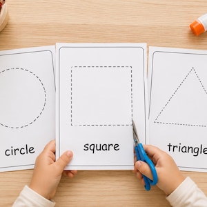May include: Three white rectangular activity sheets featuring dashed outlines of a circle, a square, and a triangle with their corresponding names printed below each shape. Blue scissors are positioned to cut along the square's dashed line.