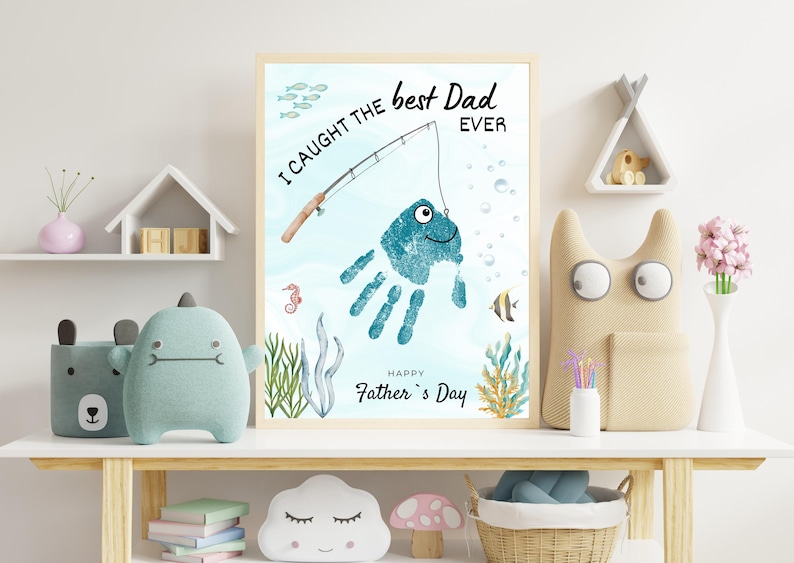 May include: A framed print featuring a teal handprint transformed into a fish with a fishing rod graphic. The text reads I CAUGHT THE best Dad EVER and HAPPY Father's Day. The background includes watercolor illustrations of seaweed, bubbles, and fish.
