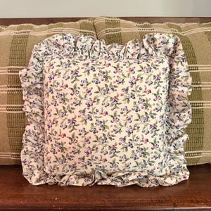 May include: A square throw pillow featuring a white background with a repeating pattern of small blue berries and green leaves. The pillow is finished with a gathered fabric ruffle border that extends around all four sides of the square cushion.