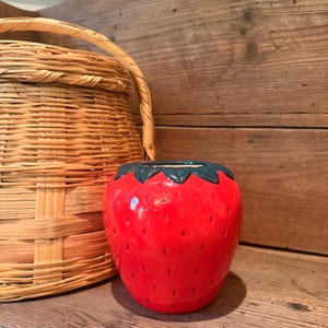 May include: A ceramic strawberry-shaped container with a green leafy top. The container is bright red with small, evenly spaced dimples. A woven basket with a handle is to the left of the container. The background is a wooden surface.