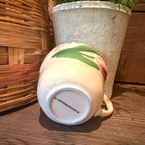 以下が含まれることがあります： 花柄と小さな取っ手が付いた、薄いピンク色のセラミックマグカップ。「by ANTHROPOLOGIE」の文字が底にあります。マグカップは木製の表面に置かれています。