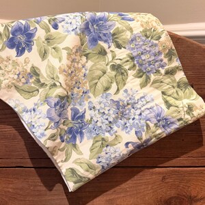 May include: A folded fabric textile featuring a floral pattern with blue and purple blossoms, green leaves, and cream-colored accents. The botanical print displays clusters of flowers and foliage arranged across the off-white background surface.