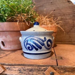 May include: A small, gray ceramic jar with a lid, featuring blue abstract swirl designs. The jar has a small blue knob on top and two small handles. The jar is sitting on a wooden surface.