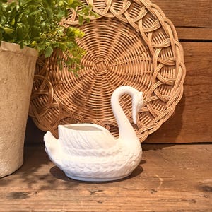 May include: A white ceramic swan planter with detailed feather textures. The swan is positioned in front of a woven wicker plate and a textured pot with greenery. The background is a wooden surface.