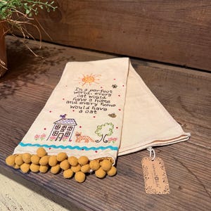 May include: A cream-colored cotton tea towel featuring embroidered designs of a house, a cat, a tree, a sun, and a bee. The text reads, In a perfect world, every cat would have a home and every home would have a cat. It has mustard yellow pom-pom trim.