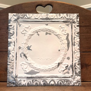May include: A square metal ceiling tile featuring a raised embossed wreath pattern in the center and a decorative border. The surface has a distressed finish with white paint peeling to reveal patches of dark gray metal underneath the texture.