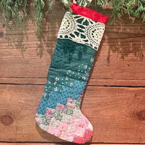 May include: A quilted Christmas stocking featuring a patchwork design of various floral and geometric fabric patterns in shades of green, blue, and pink. The top cuff is accented with a cream-colored crocheted lace trim and a bright red fabric border.