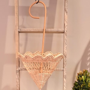May include: A light brown, woven wicker umbrella-shaped basket with a scalloped edge, hanging from a wooden handle. The handle curves into a hook, suspended by a black cord. The basket is hanging on a wooden ladder.