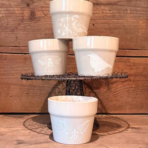 May include: Four small, off-white ceramic flower pots with embossed nature designs. The designs include a bird, a bee, a bird, and a butterfly. The pots are displayed on a decorative wire stand and wooden surface.
