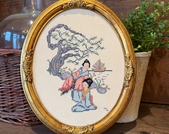 May include: An oval gold-toned frame containing a cross-stitch embroidery on white fabric. The design features two figures in traditional clothing beneath a stylized tree, with a small sailboat on the water in the background.