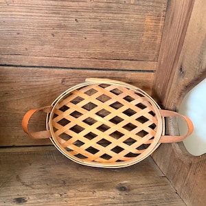 May include: Oval-shaped woven basket with a lattice design on the lid. The basket has two brown leather handles. The basket is made of light-colored wood.