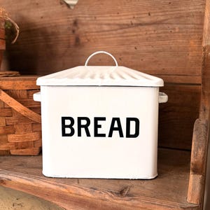 May include: A white rectangular bread box with a lid and handle, the word "BREAD" is printed in black on the front. The bread box is sitting on a wooden shelf, with a wooden bench in the background.