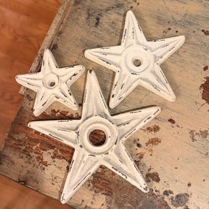May include: Three off-white, five-pointed star-shaped metal objects with a distressed finish. Each star features a central circular opening and raised ridges extending from the center to each point, showing dark metal underneath the paint.