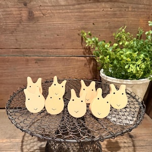 May include: A collection of six light yellow wooden bunny-shaped ornaments with black dot eyes and a curved mouth. The bunnies are arranged on a vintage wire cake stand. The background features a wooden wall and a potted plant.