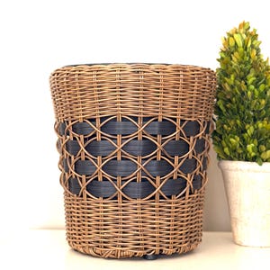 May include: A round, woven wicker basket with a dark blue geometric pattern around the middle. The basket is brown and has a sturdy, cylindrical shape. A small, green plant in a white pot is to the right.