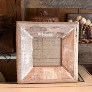 May include: A square wooden picture frame with a light brown finish and white distressed paint accents. The frame holds a piece of tan paper featuring dark brown horizontal lines and handwritten cursive script text in dark ink.