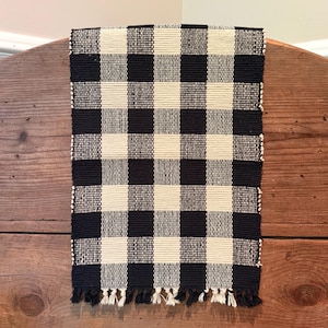 May include: A black and cream checkered table runner with fringe. The runner is made of woven fabric and features a classic buffalo check pattern. The neutral colors and simple design make it suitable for various decor styles.