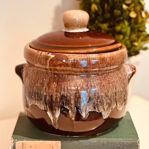 May include: A brown ceramic casserole dish with a lid and two side handles. The dish has a dripped glaze effect with lighter brown and white accents. It sits atop a vintage green book.