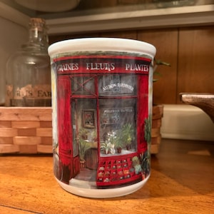 May include: A white ceramic mug featuring a detailed illustration of a red storefront with the text GRAINES FLEURS PLANTES, AU BON JARDINIER, and MARIEROSE DUFFEL. The design depicts a shop entrance with potted plants and small seed displays.