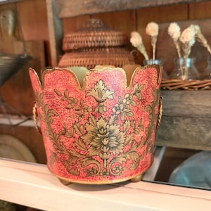 May include: A cylindrical metal planter with a scalloped top edge and three small feet. The exterior features a textured coral-red finish with a prominent gold-toned floral and leaf pattern. Two twisted gold-toned metal handles are attached to the sides.