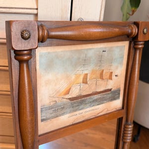 May include: Wooden framed mirror featuring a vintage print of the clipper ship "Nightingale." The frame is a warm brown with decorative accents, including a floral design on the top corners. The print shows the ship sailing on the water.