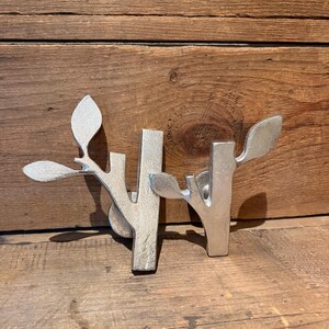 May include: Two silver-colored metal wall hooks, each shaped like a stylized tree branch with a single leaf extending from the top. The hooks feature a textured, matte finish and a vertical post design, providing a functional and decorative element.