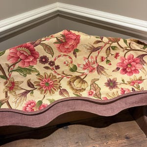May include: A fabric valance featuring a cream background with a floral pattern of pink, red, and green blossoms and leaves. The bottom edge is scalloped and finished with a solid dark red, finely textured border trim along the entire length.