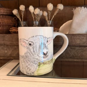 May include: A white ceramic mug featuring a hand-painted illustration of a grey lamb with pink-tinted ears. The lamb is positioned in front of a green fence. The mug has a curved handle on the right side and a smooth, glossy finish throughout.