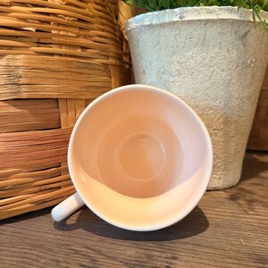 以下が含まれることがあります： 白い取っ手が付いた、明るいピーチ色のティーカップが木製の表面に置かれています。カップは手前にあり、背景には編みかご、小さな鉢植えの植物があります。カップの内側はソフトピーチ色です。