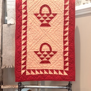 May include: A rectangular quilted table runner featuring a cream-colored center panel with two red basket motifs. The border consists of a red and cream sawtooth pattern. The entire piece is finished with a red fabric edge and quilted texture.