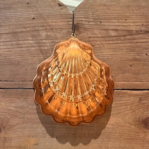 May include: A shiny, metallic copper-colored mold shaped like a scallop shell. The mold features a detailed, ribbed surface with a scalloped edge and a small metal loop at the top for hanging, reflecting light across its curved, reflective surface.