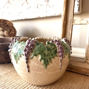 May include: A beige ceramic bowl with a textured surface, adorned with green leaves and purple grape clusters around the rim. The bowl is likely handmade and adds a rustic touch to home decor.