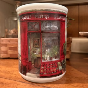 May include: A white ceramic mug featuring a detailed illustration of a red storefront with the text GRAINES FLEURS PLANTES, AU BON JARDINIER, and MARIEROSE DUFFELL. The shop front displays various potted plants and a display of seeds.