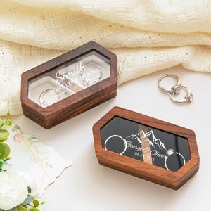 May include: Two wooden ring boxes with clear tops, one with the names "Smith, Ronald & Donna" and the other with "George & Oliver" and a mountain graphic. Two silver rings with stones are next to the boxes.