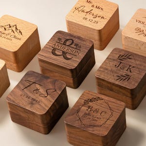 Può includere: Una collezione di scatole di legno personalizzate, alcune chiare e altre scure, con nomi e date incisi. Le scatole presentano vari disegni, tra cui silhouette di montagne, motivi floreali e iniziali. I testi includono nomi come "Anderson" e date.