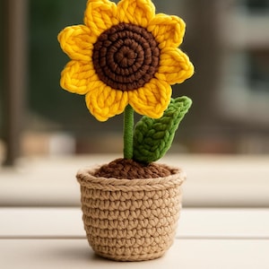 Può includere: Un girasole all'uncinetto fatto a mano in un piccolo vaso. Il girasole ha petali gialli e un centro marrone. Anche il gambo e la foglia verdi sono all'uncinetto. Il vaso è di colore marrone chiaro.