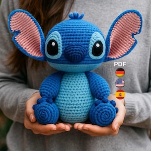 Puede incluir: Un peluche azul tejido a mano con grandes ojos negros y grandes orejas con el interior rosa. El juguete está sostenido en las manos de una persona. El juguete es un personaje de una popular película de animación.