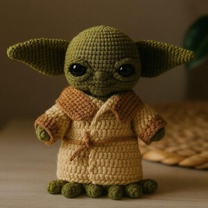 May include: A handmade crochet doll of a character with large ears and big black eyes. The doll is wearing a tan and brown robe. The doll's skin is green, and it has small green feet. The doll is standing on a wooden surface.