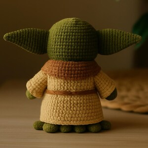 May include: A handmade crochet figure of a character with large ears, wearing a tan robe with a brown collar and belt. The figure is green and tan, with small green feet. The figure is a popular character from a science fiction franchise.