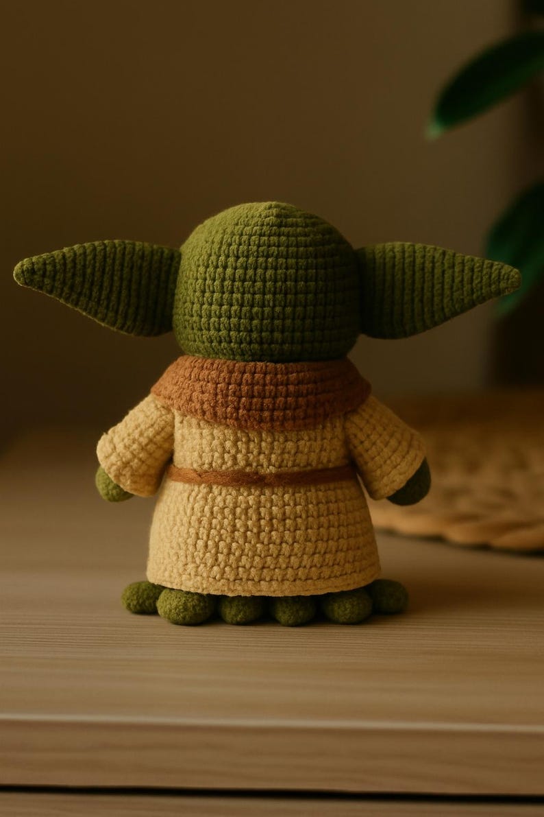 May include: A handmade crochet figure of a character with large ears, wearing a tan robe with a brown collar and belt. The figure is green and tan, with small green feet. The figure is a popular character from a science fiction franchise.