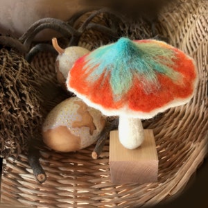 Puede incluir: Un hongo hecho a mano de fieltro con una base blanca y una tapa colorida. La tapa es naranja, roja y verde con un centro azul. El hongo está sentado sobre una base de madera en una cesta de mimbre.