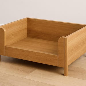 May include: A rectangular wooden pet bed with a light brown finish. The bed has a low profile with a raised back and sides, supported by four short legs. The design is simple and modern.