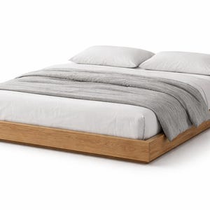 以下が含まれることがあります： 明るい木製フレームのモダンなプラットフォームベッド。白いシーツ、2つの白い枕、灰色の毛布が敷かれています。ベッドフレームの長さは約200cm、幅は約152cmです。